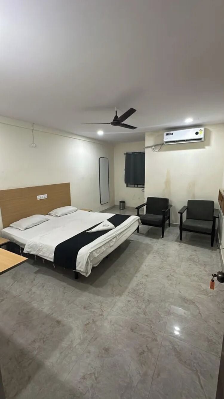 A hotel room with a bed, two chairs, a ceiling fan, and an air conditioning unit.
