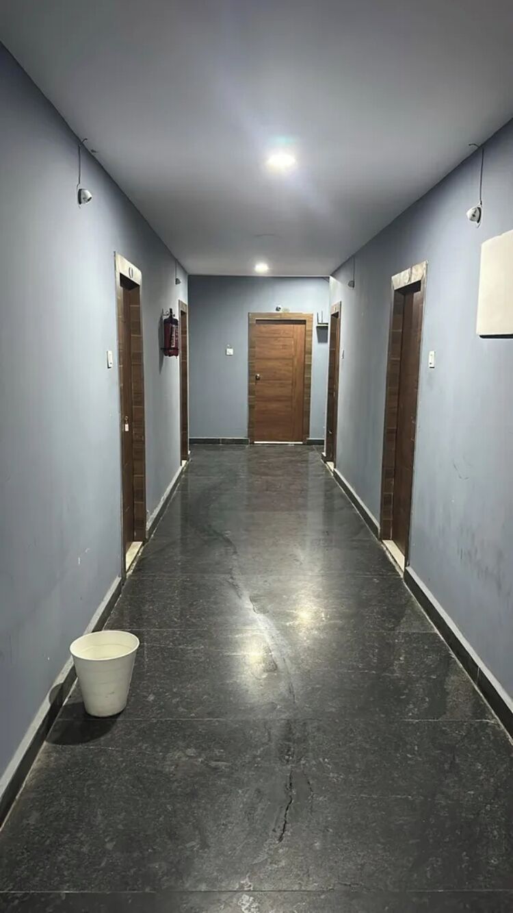 Un pasillo con una puerta de madera al final, un cubo blanco en el suelo y un accesorio de iluminación en el techo.