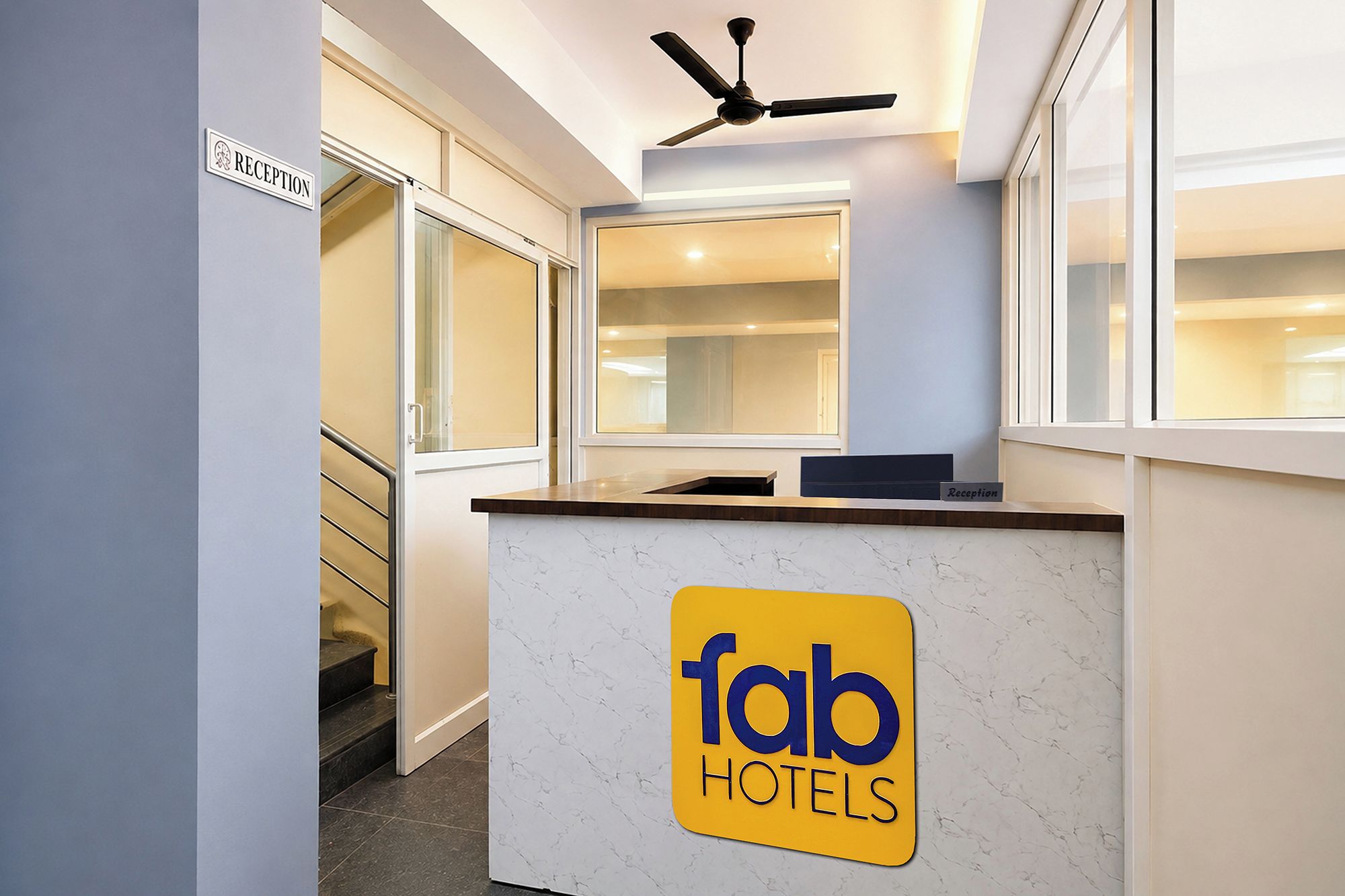 Ein Empfangsschalter mit einem Schild für 'fab hotels', ein Deckenventilator und eine Treppe, die in einen anderen Bereich führt.