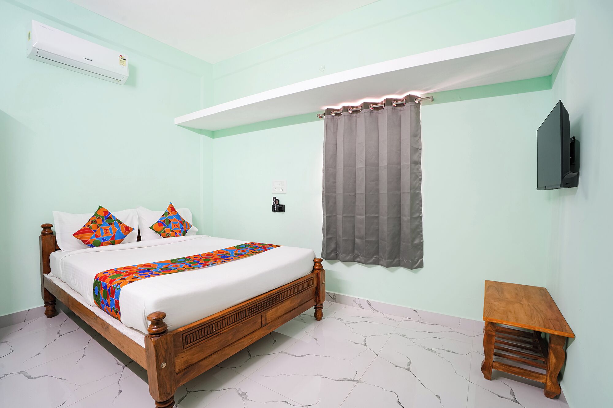 A bedroom with a bed, a bedside table, a TV mounted on the wall, and a window with curtains.