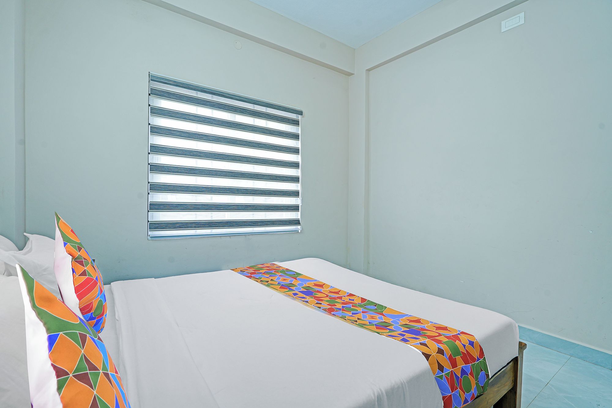 A single bed with a colorful bedspread in a room with light blue walls and a window with horizontal blinds.