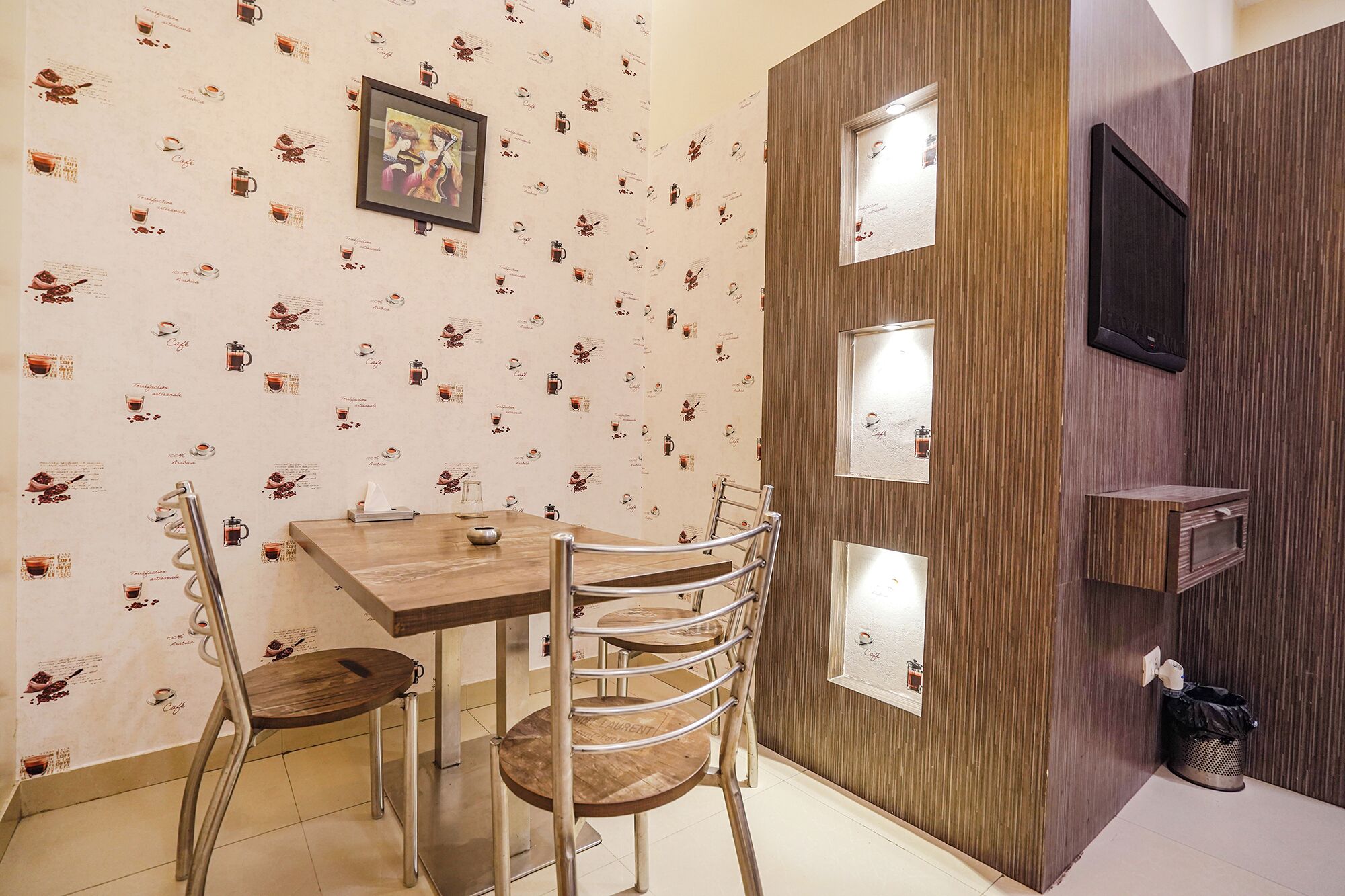 A dining area with a wooden table and chairs, a wall with a coffee-themed wallpaper, and a mounted TV.