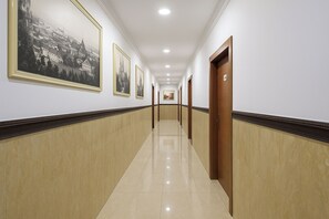 A long, well-lit hallway with framed black and white photographs on the walls, wooden doors, and a tiled floor.