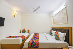 A hotel room with two single beds, a large bed, a ceiling fan, a wall-mounted TV, and a framed artwork.