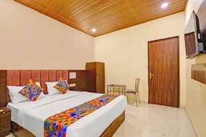 A hotel room with a wooden ceiling, a bed with colorful bedding, a wooden wardrobe, a small table, and a door. - Via Surya Grand (Mumbai)
