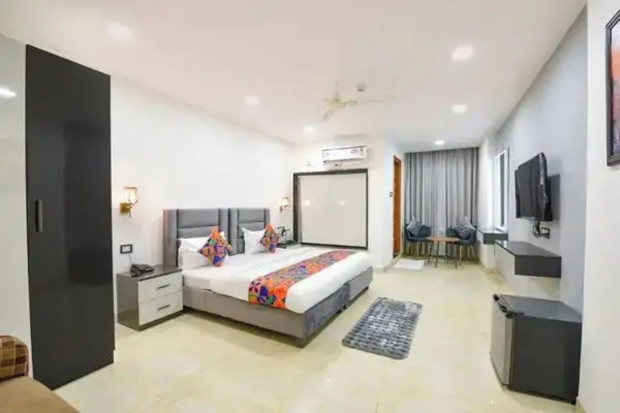A modern hotel room with a bed, bedside tables, a desk, a chair, a TV, and an air conditioning unit.