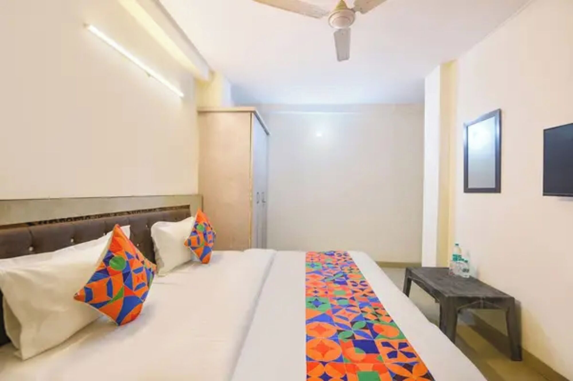 A hotel room with a bed, pillows, a bedside table, a ceiling fan, and a wall-mounted TV.