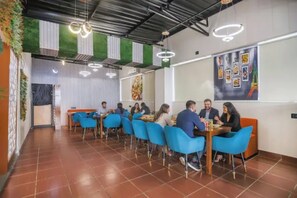 A dining area with blue chairs and tables, a green wall with vertical blinds, and a ceiling with exposed beams and circular light fixtures.