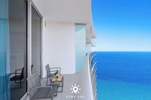 Ein Balkon mit Glasgeländer und Blick auf den Ozean, ein Tisch mit Stühlen und eine kleine Pflanze.