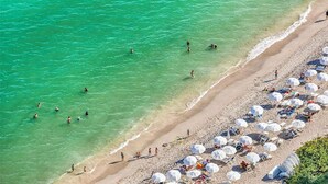 Luftaufnahme eines Strandes mit klarem türkisblauem Wasser, Menschen beim Schwimmen und Spazierengehen am Ufer sowie zahlreichen Strandsonnenschirmen und Liegestühlen.