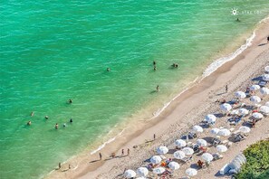 Luftaufnahme eines Strandes mit klarem türkisblauem Wasser, Menschen beim Schwimmen und Spazierengehen am Ufer sowie zahlreichen Strandsonnenschirmen und Liegestühlen.