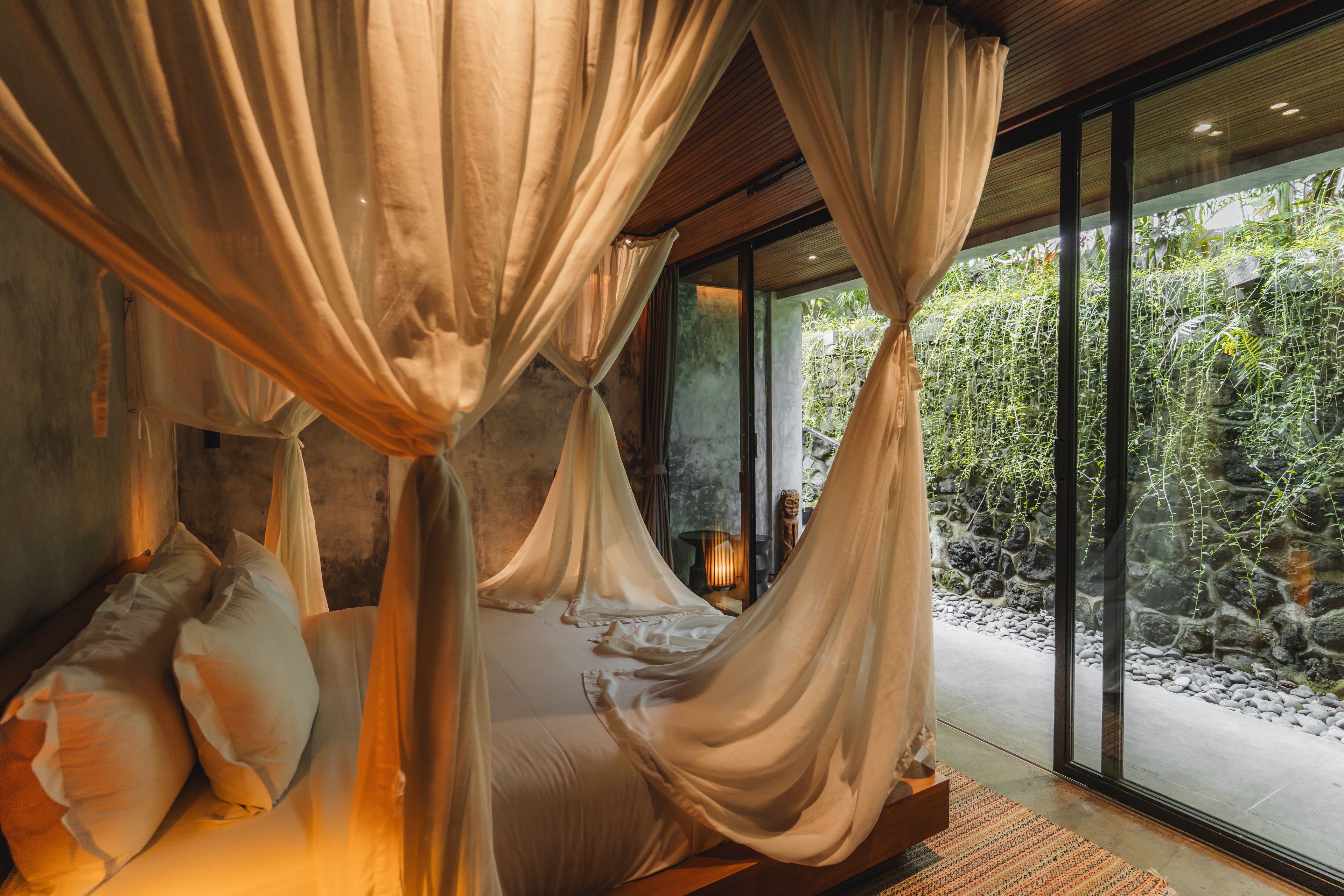 A room with a bed under a canopy, a stone wall, and a view of greenery outside.