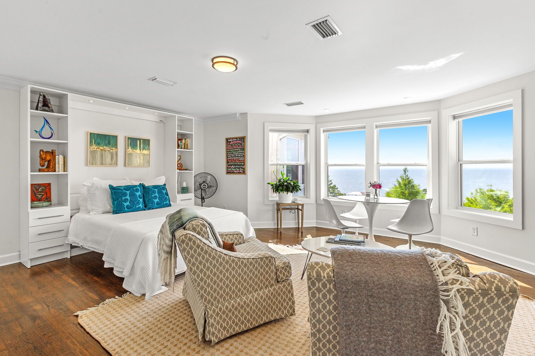 A spacious bedroom with a large bed, a sitting area with a table and chairs, a bookshelf, and a view of the ocean through large windows.