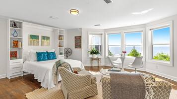 A spacious bedroom with a large bed, a sitting area with a table and chairs, a bookshelf, and a view of the ocean through large windows.