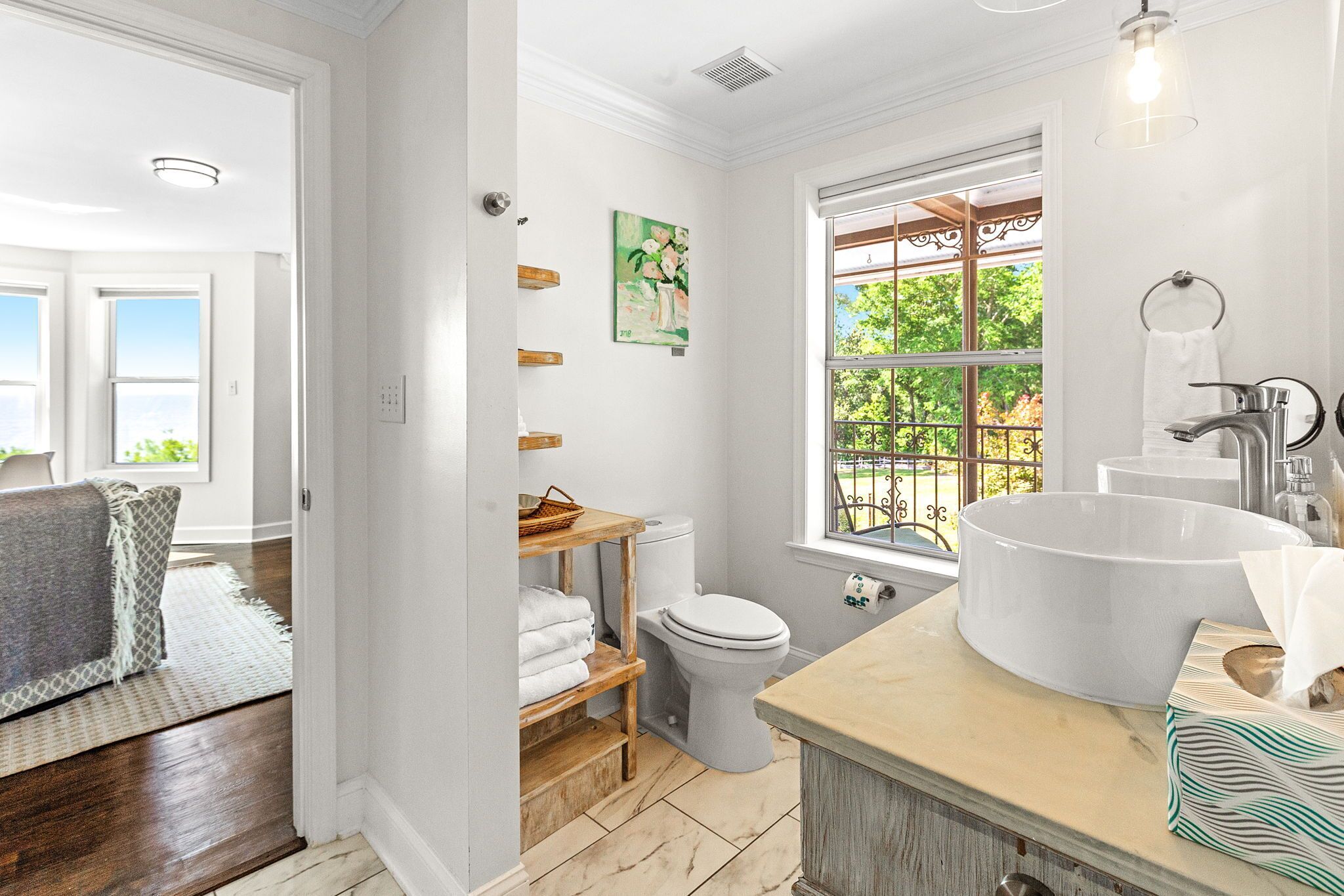 A bathroom with a large sink, toilet, and window. There is a towel rack and a painting on the wall.