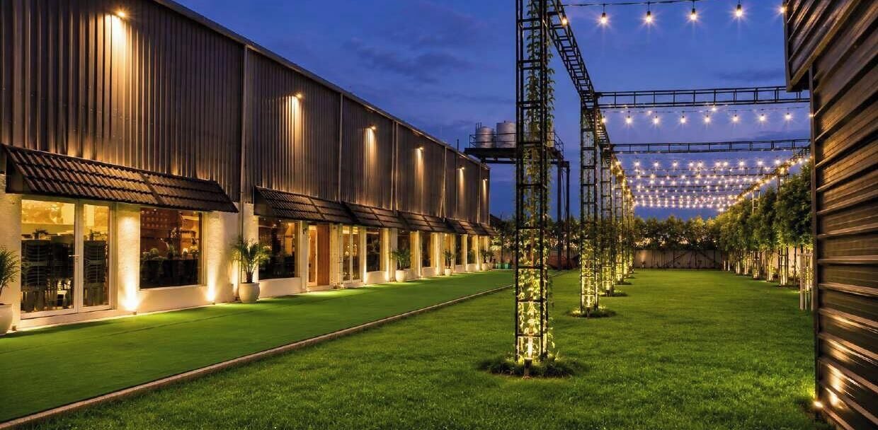 Un bâtiment à la structure longue et étroite, avec un espace verdoyant devant et des guirlandes lumineuses suspendues au-dessus.