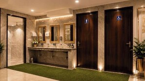 Baño de hotel con paredes de mármol, un lavamanos central con espejos y dos puertas, una de ellas con el símbolo de accesibilidad para sillas de ruedas.