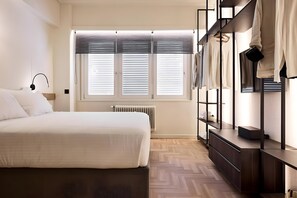 A bedroom with a bed, a radiator, a window with blinds, a shelf with clothes, and a desk. - Estia Athens – Your Luxurious Urban Retreat (Athens)