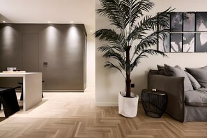 A modern living room with a wooden herringbone floor, a white potted plant, a grey sofa, and a black side table. - Estia Athens – Your Luxurious Urban Retreat (Athens)