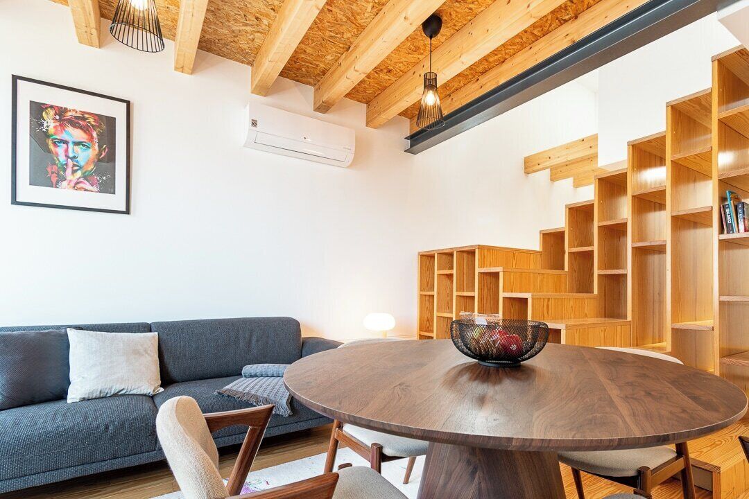 A modern living room with a round wooden table, a gray sofa, and a wooden bookshelf.