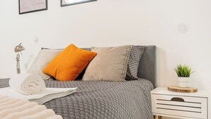 Uma cama bem arrumada com um edredom cinza estampado, uma toalha branca e um único travesseiro laranja. Há uma mesa de cabeceira branca com uma planta em cima.