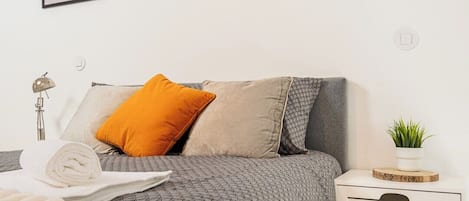 A neatly made bed with a grey patterned bedspread, a white towel, and a single orange pillow. There is a white bedside table with a plant on top.