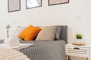 Ein ordentlich bezogenes Bett mit einer grauen, gemusterten Tagesdecke, einem weißen Handtuch und einem einzelnen orangefarbenen Kissen. Ein weißer Nachttisch mit einer Pflanze darauf.