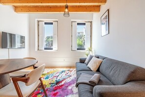 A modern living room with a gray sofa, a wooden dining table, and a colorful rug. There are two windows with white frames, a ceiling light, and a flat-screen TV mounted on the wall. - Modern Apt in Historic Porto's Largo da Lapa (Porto)