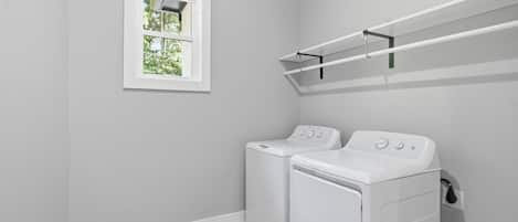 A laundry room with two white washing machines, a window with a view of greenery, and a metal shelf above the machines.