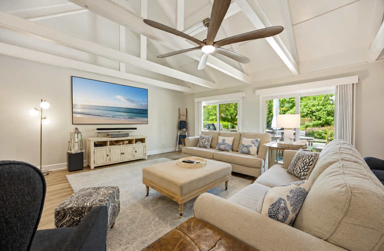 A spacious living room with a ceiling fan, a large flat-screen TV, a beige sofa set, and a view of greenery outside.