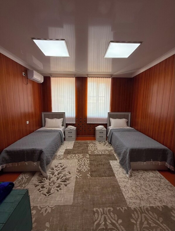 A room with two beds, a large window with blinds, an air conditioning unit, and a patterned rug.