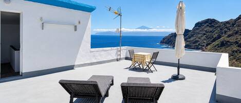 A rooftop terrace with two lounge chairs, a table, and an umbrella, overlooking a coastal landscape with a mountain in the distance.