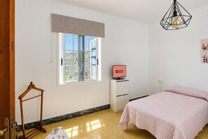 Ein Zimmer mit einem Bett, einem Fernseher auf einem Schrank, einem Fenster mit Jalousien und einer Hängelampe.