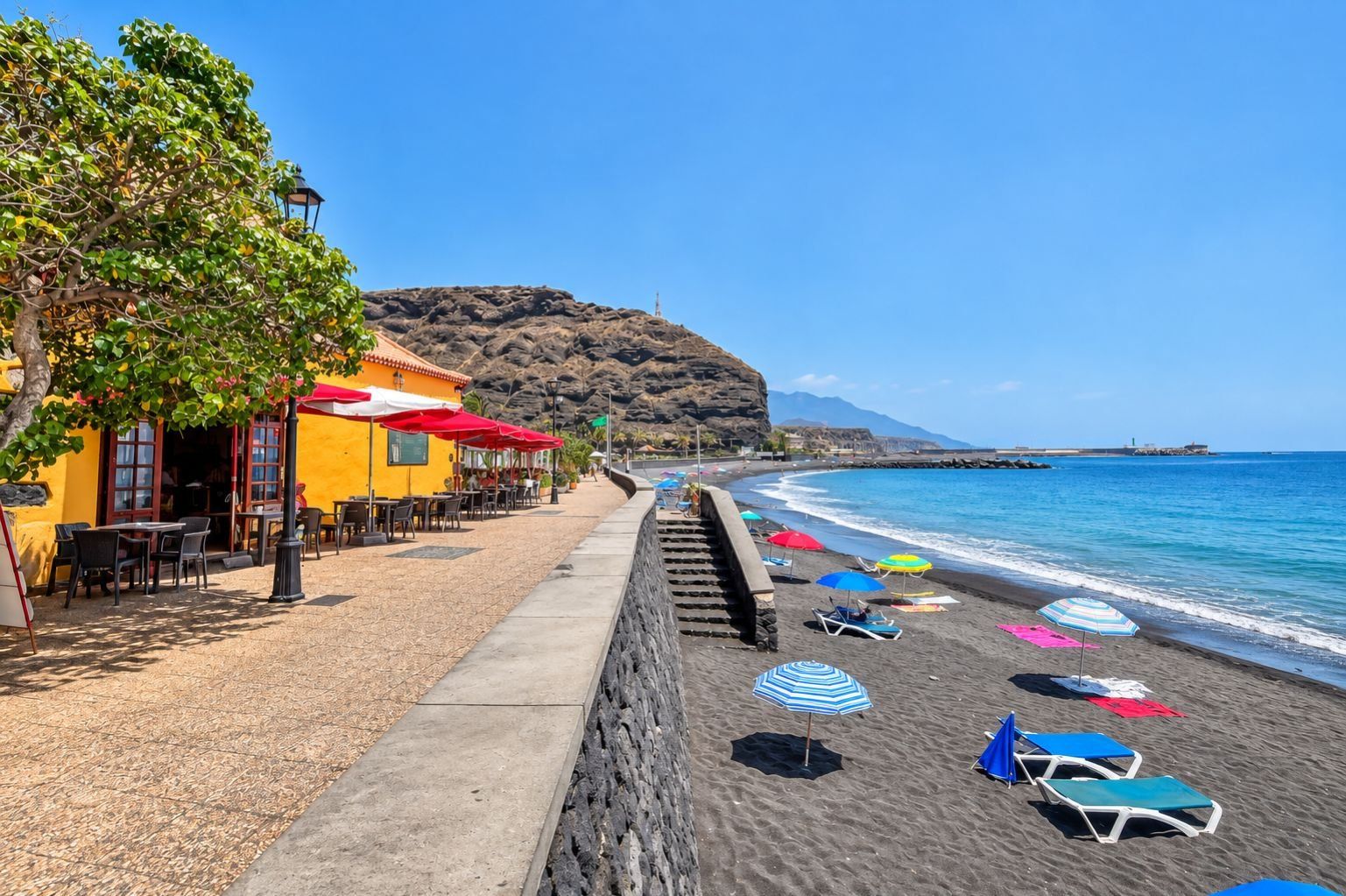 A coastal beach scene with a row of outdoor seating and umbrellas, a stone wall, and a clear view of the sea and distant mountains.