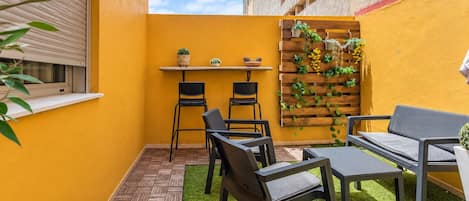 Une petite terrasse extérieure avec une table et des chaises, un jardinier en bois avec des plantes et une fenêtre avec des stores.