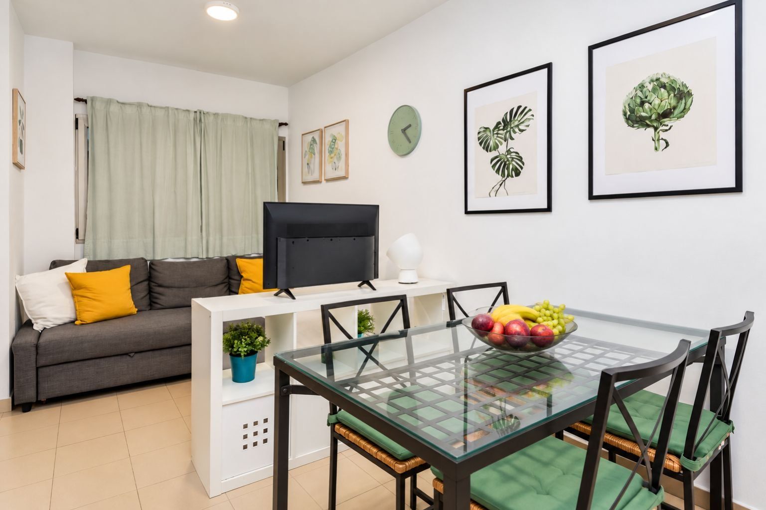 A modern living room with a sofa, a flat-screen TV, a glass dining table with chairs, and artwork on the walls.