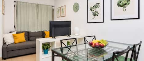 A modern living room with a sofa, a flat-screen TV, a glass dining table with chairs, and artwork on the walls.