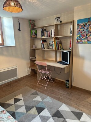 Ein Zimmer mit Schreibtisch aus Holz, einem rosa Stuhl, einem Bücherregal und einem gemusterten Teppich.