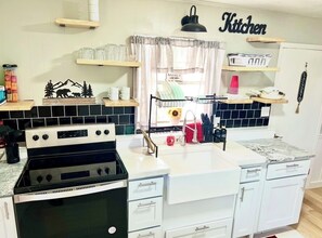 A kitchen with a stove, sink, and white cabinets. There are shelves above the counter and a sign that reads 'Kitchen'.