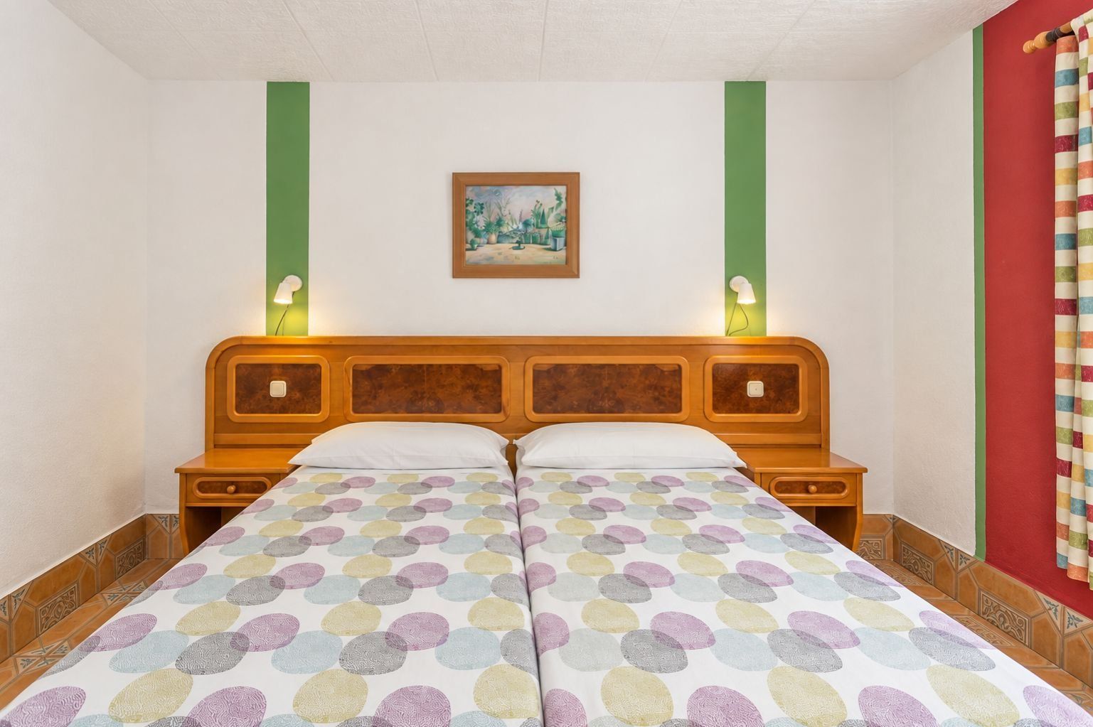 Un letto matrimoniale con copripiumino a pois, due comodini con lampade, un quadro incorniciato appeso al muro e una tenda rossa sulla destra.