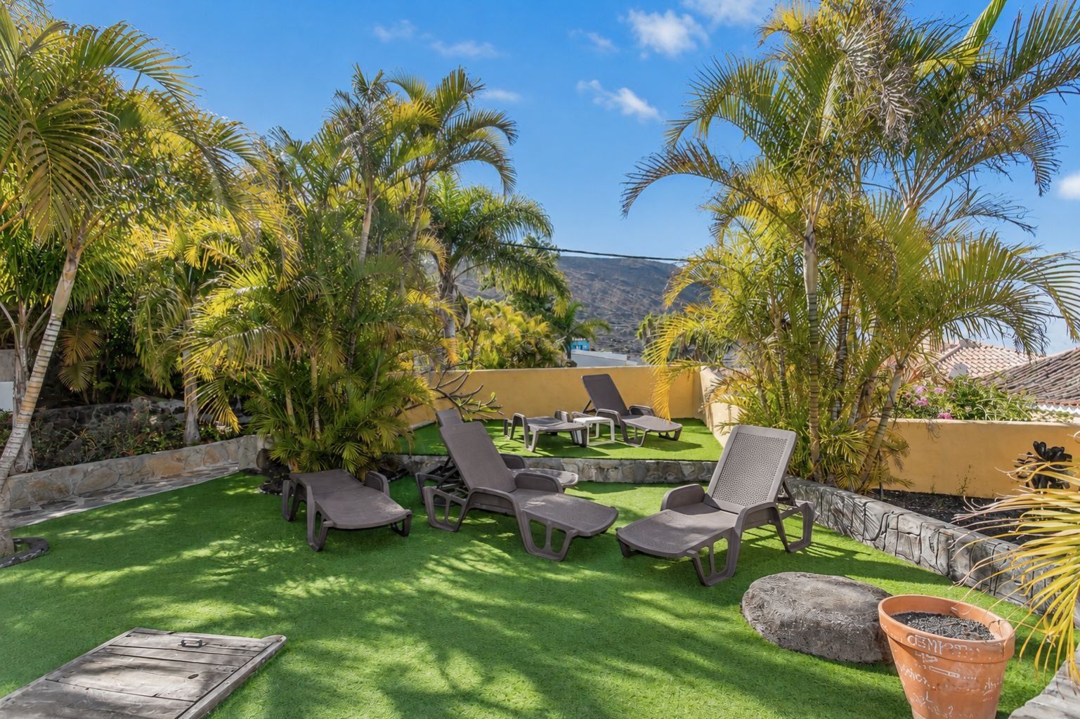 Un área de estar al aire libre con sillones reclinables y césped artificial verde, rodeada de palmeras y un muro de piedra.