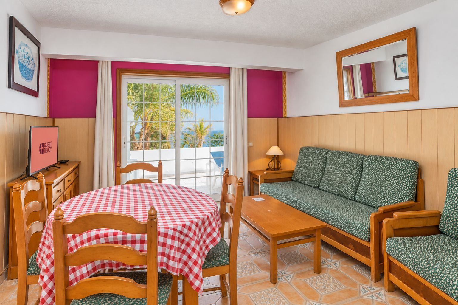 Una habitación con una mesa a cuadros, un sofá, un televisor y una ventana con vista a palmeras.