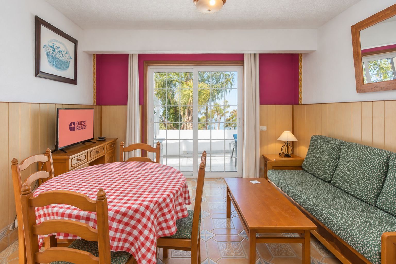 Una habitación con una mesa a cuadros, un sofá, un televisor y vistas a palmeras a través de la ventana.