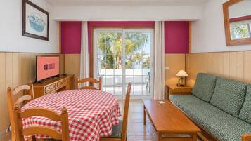 Una habitación con una mesa a cuadros, un sofá, un televisor y vistas a palmeras a través de la ventana.