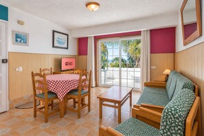 Ein Zimmer mit Esstisch, Stühlen, einer Couch, einem Fernseher und Blick ins Freie.