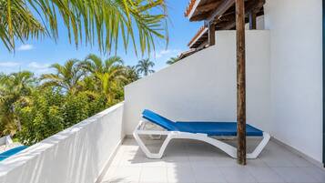 A sunlit balcony with a white tiled floor, a blue lounge chair, and a view of palm trees and a clear sky.