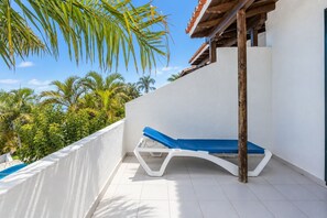 Ein sonniger Balkon mit gefliestem Boden, einem blauen Liegestuhl und Blick auf Palmen und einen klaren Himmel.