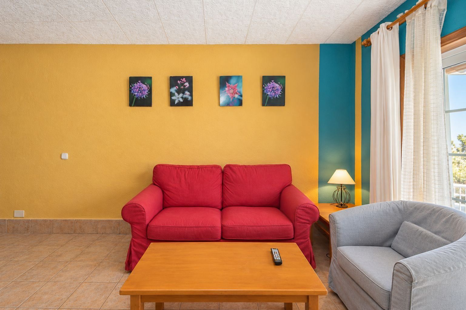 A living room with a red sofa, a grey armchair, a wooden coffee table, and four framed floral paintings on the wall.