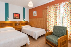 Quarto de hotel com duas camas, uma cabeceira de madeira, uma poltrona verde, cortina xadrez colorida e uma parede com uma pintura de pôr do sol.