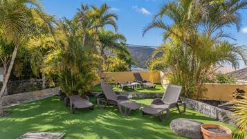 Un área de estar al aire libre con sillones reclinables y césped artificial verde, rodeada de palmeras y un muro de piedra.